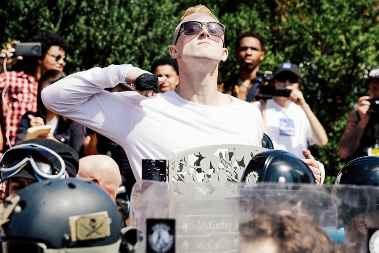 A blonde man wearing black sunglasses and a white shirt makes a throat-slash gesture with a hand that is taped as if to prepare for fistfighting.